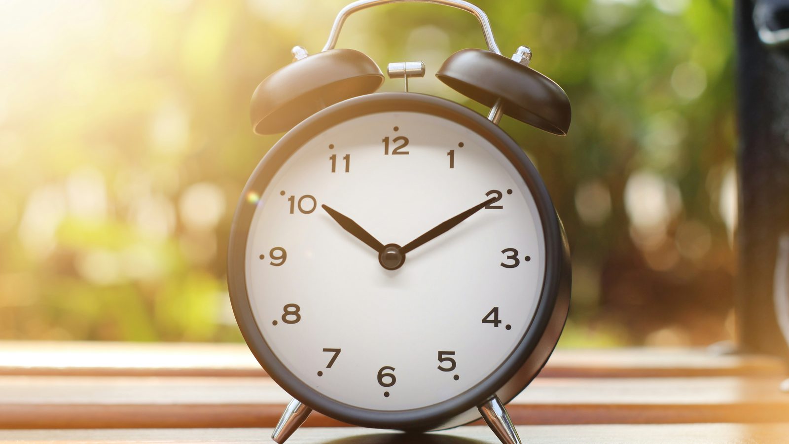 A brown alarm clock on a wooden table in warm morning light, with green foliage blurred behind it.