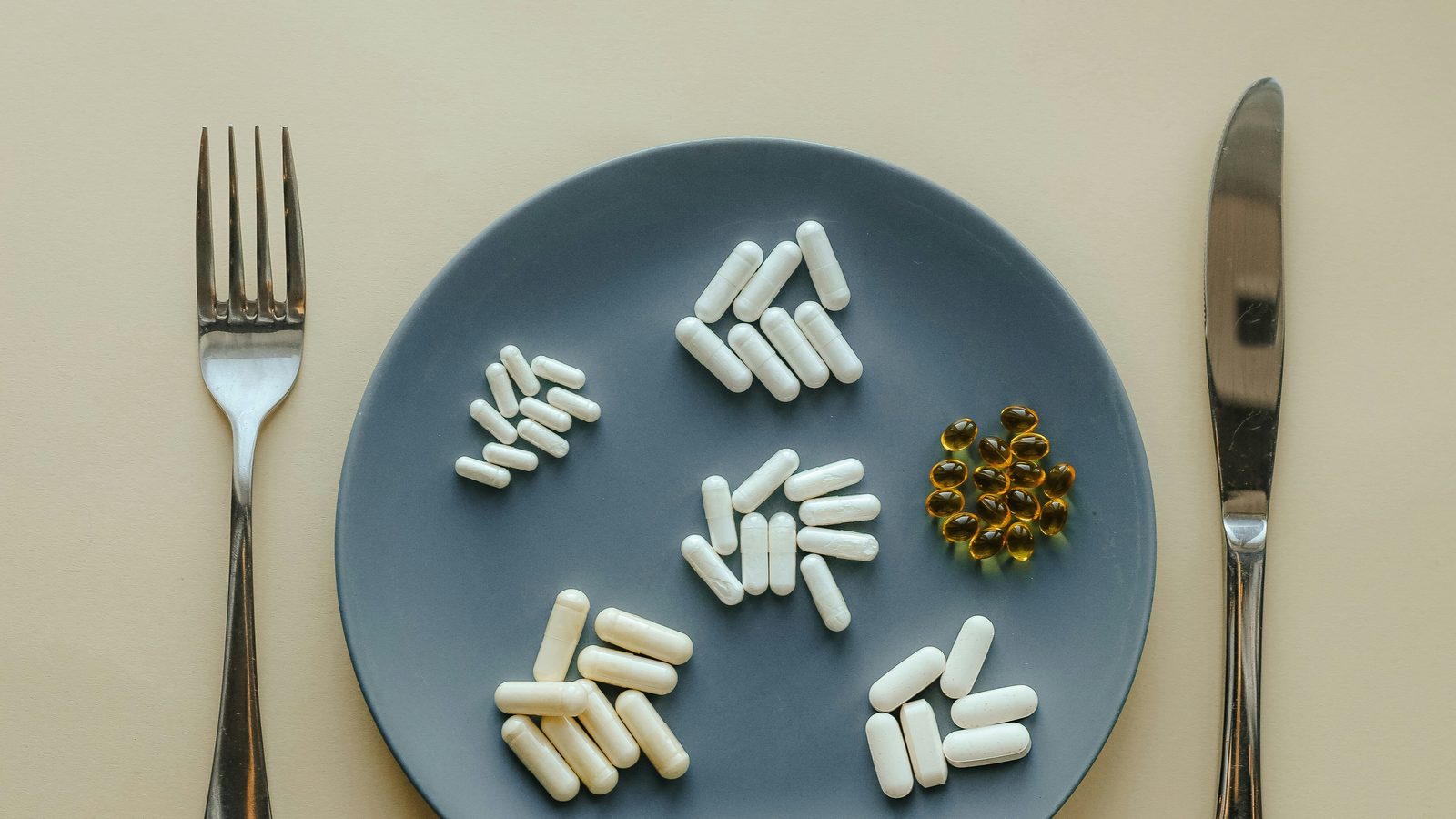 Capsules and softgels arranged in small groups on a slate-blue plate, with a fork and knife on either side.